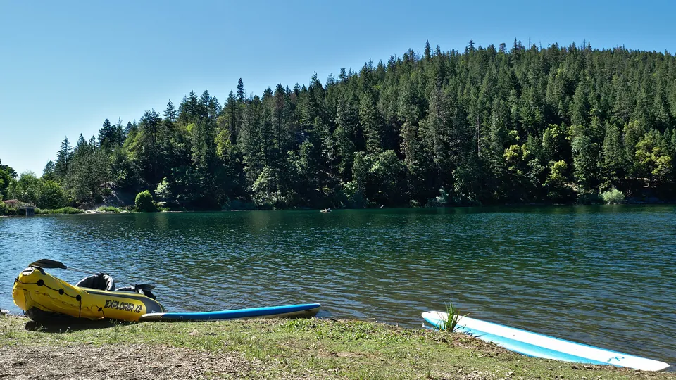Ein SUP-Board liegt am bewaldeten Seeufer, ein weiteres auf dem Wasser; daneben liegt ein gelbes Schlauchboot.